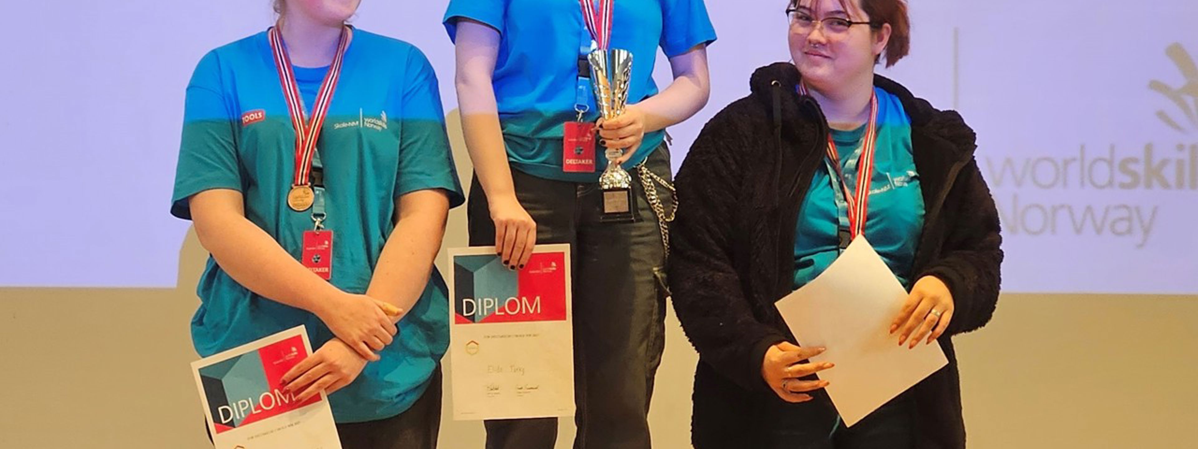 Tre jenter på et podium med diplom og medalje