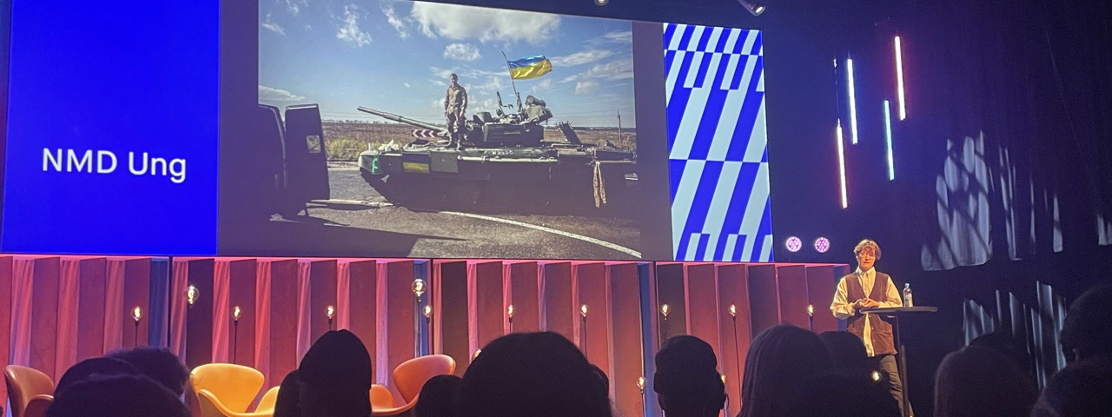 Publikum ser på presentatør på scenen med skjerm i bakgrunnen med stridsvogn fra Ukraina.